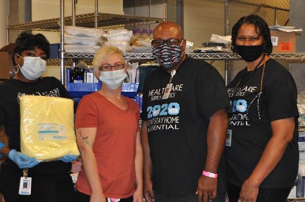 Supply Chain Technicians Temetrice Wilder (holding an isolation gown package), Sherry Sherron, Woody Johnson and Sharon Binion stand near a PAR level cart they are building for a new COVID-19 unit.